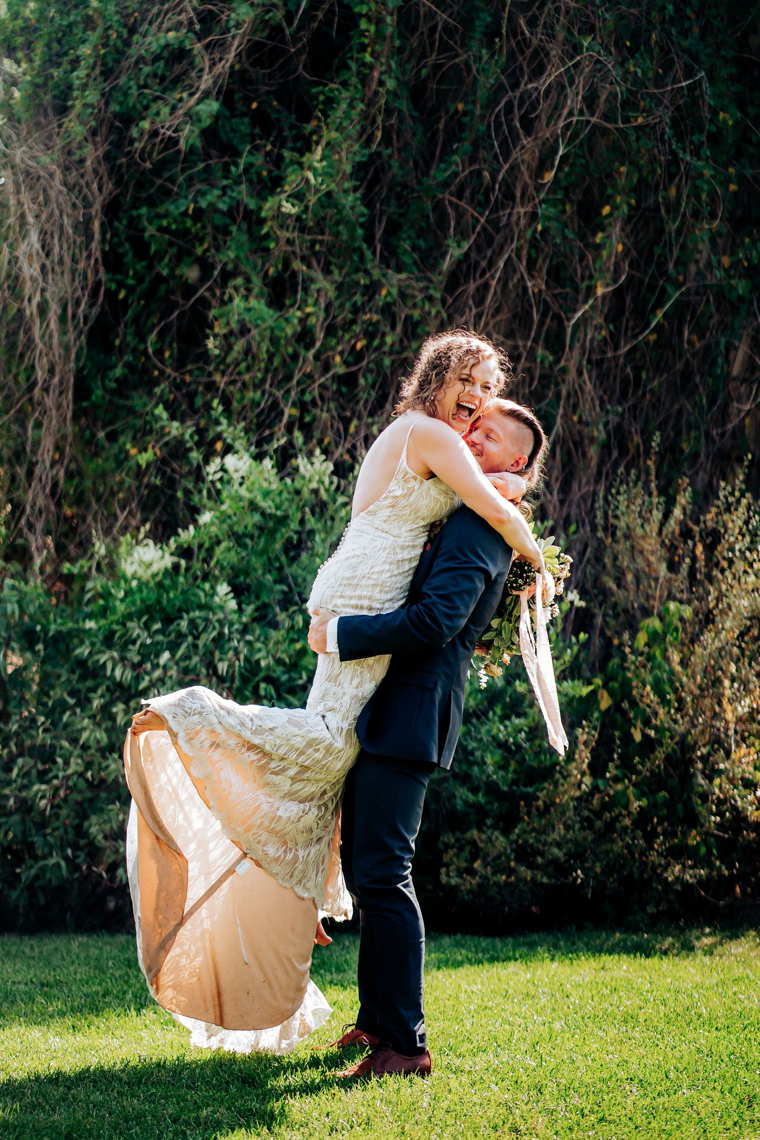 A playful wedding photo showing a couple in a celebratory lift pose on a green lawn.
