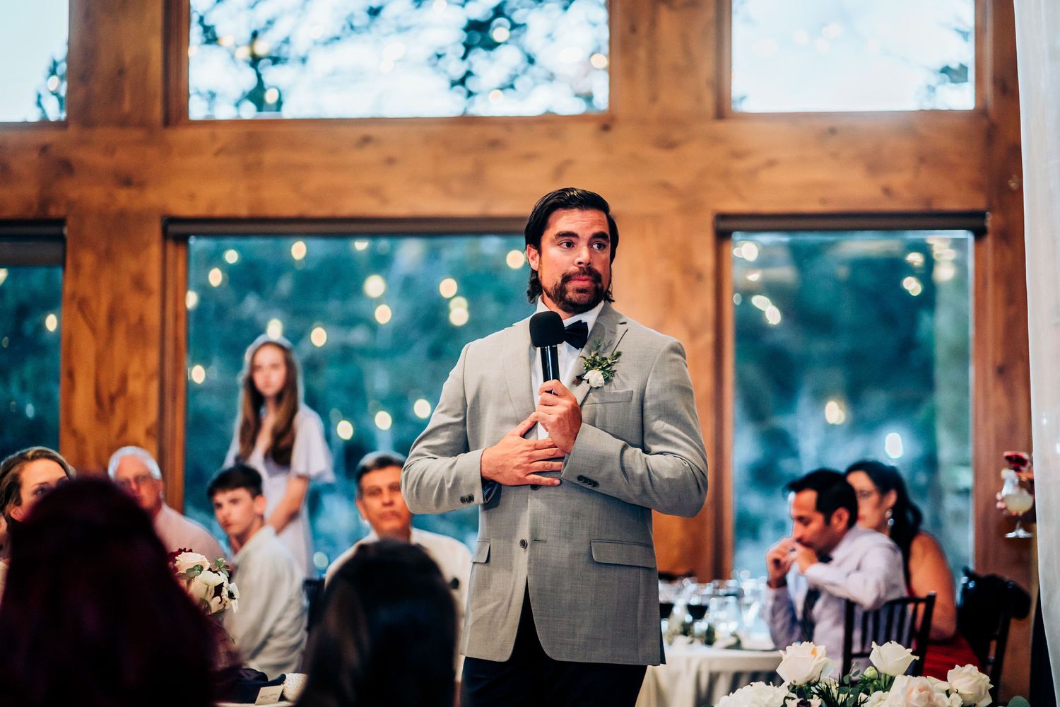 Person in grey suit giving speech at wedding reception with large wooden windows in background.