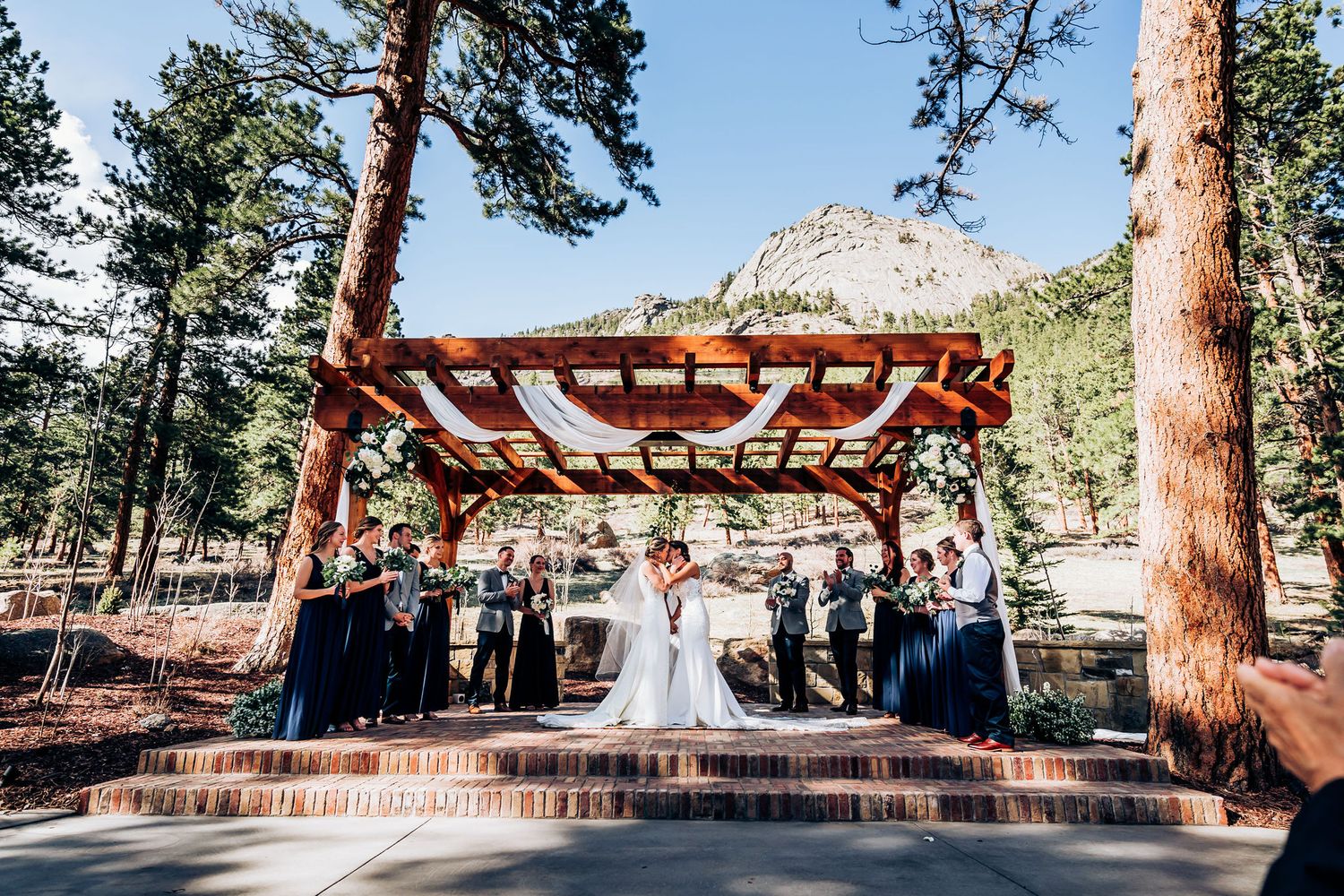 A wedding ceremony moment captured under a wooden pergola with mountains and pine trees in the background.