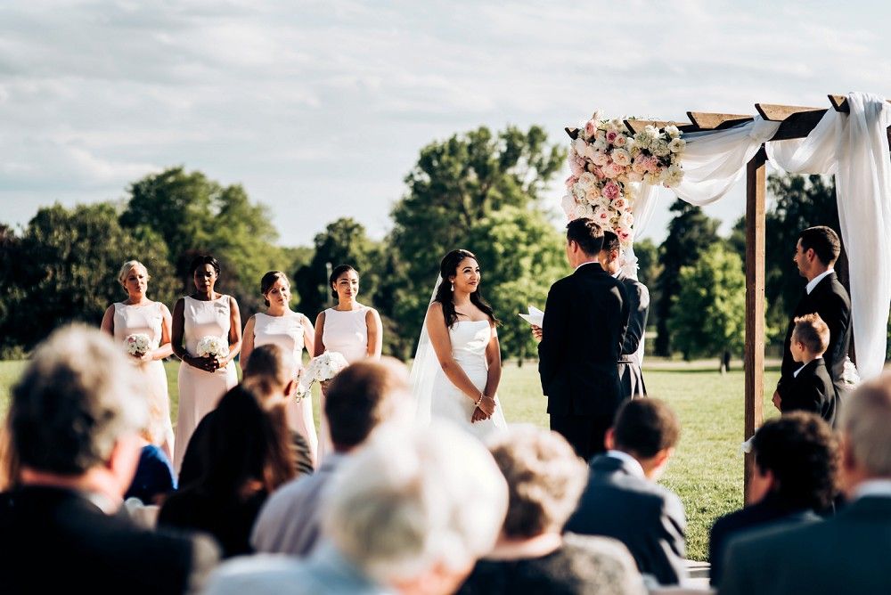 Outdoor wedding ceremony under a floral arch with guests seated on white chairs.