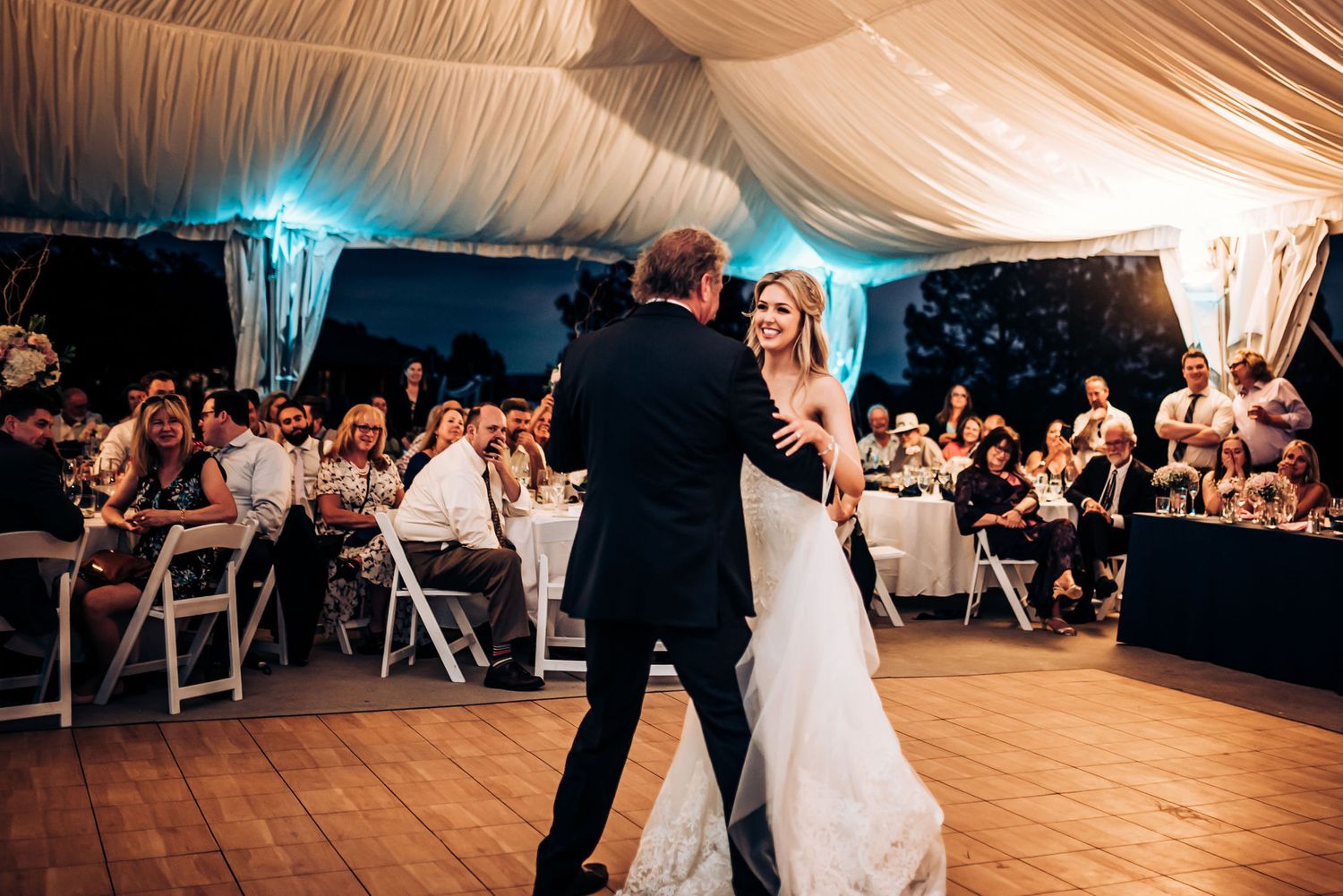 Wedding couple dances while guests watch from white folding chairs in an elegant tent reception.
