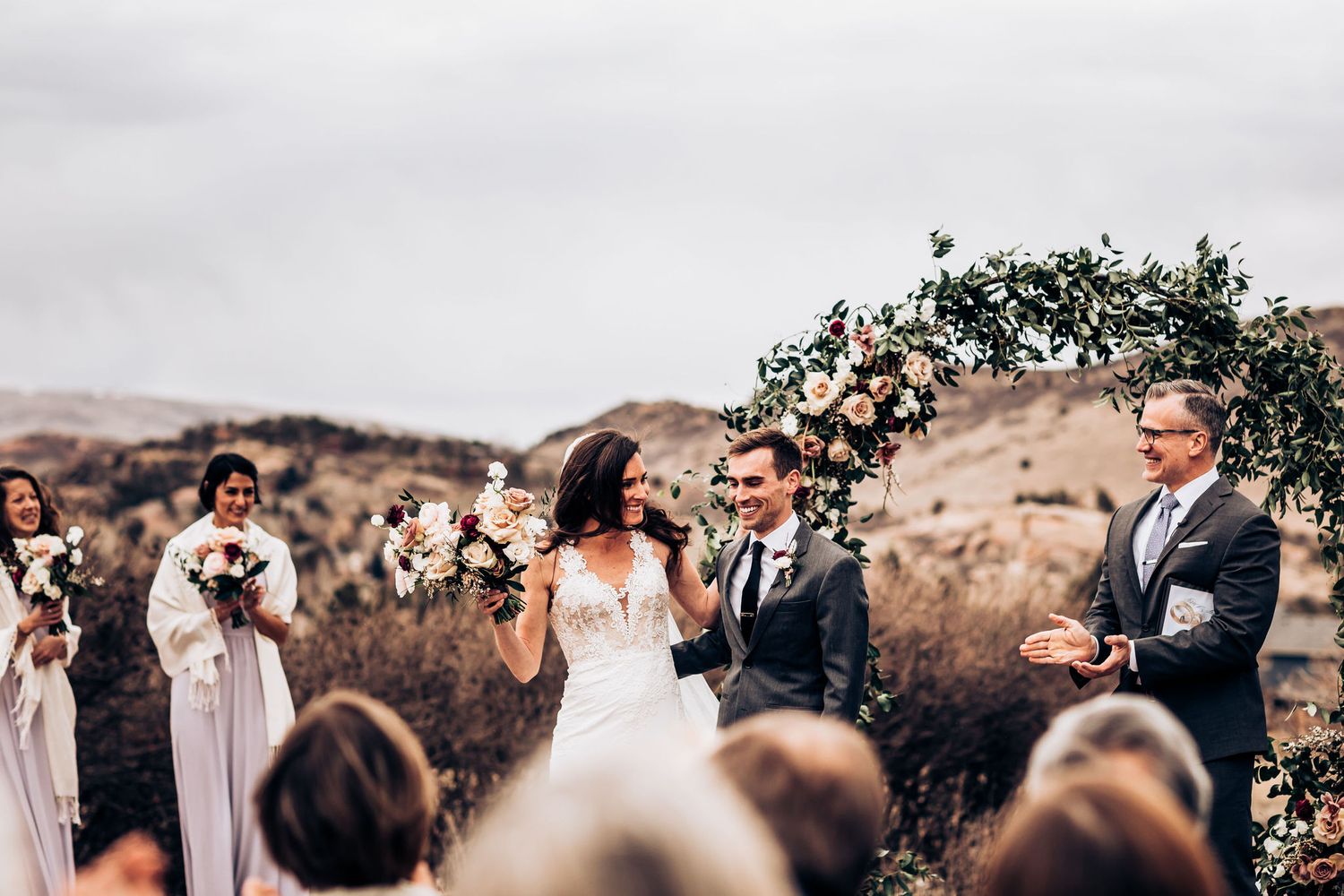 Outdoor wedding ceremony in a mountain landscape with a decorative floral arch and gathered guests.