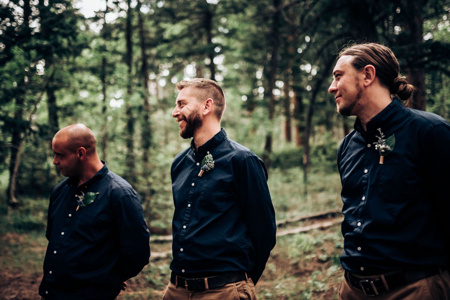 Groomsmen in coordinated navy shirts stand attentively during an outdoor forest ceremony.