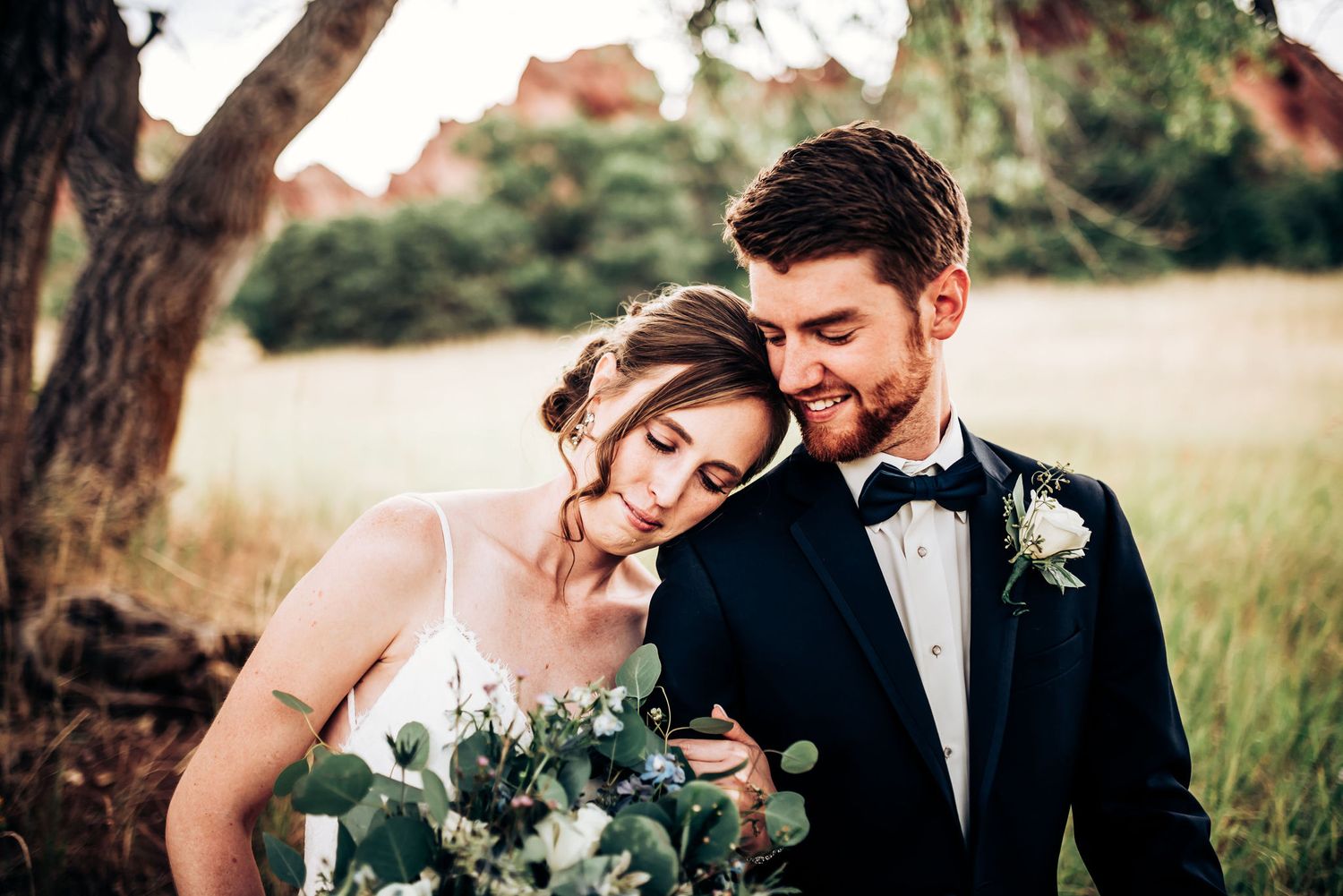 Romantic wedding couple embraces in golden field at sunset with bride holding eucalyptus bouquet.
