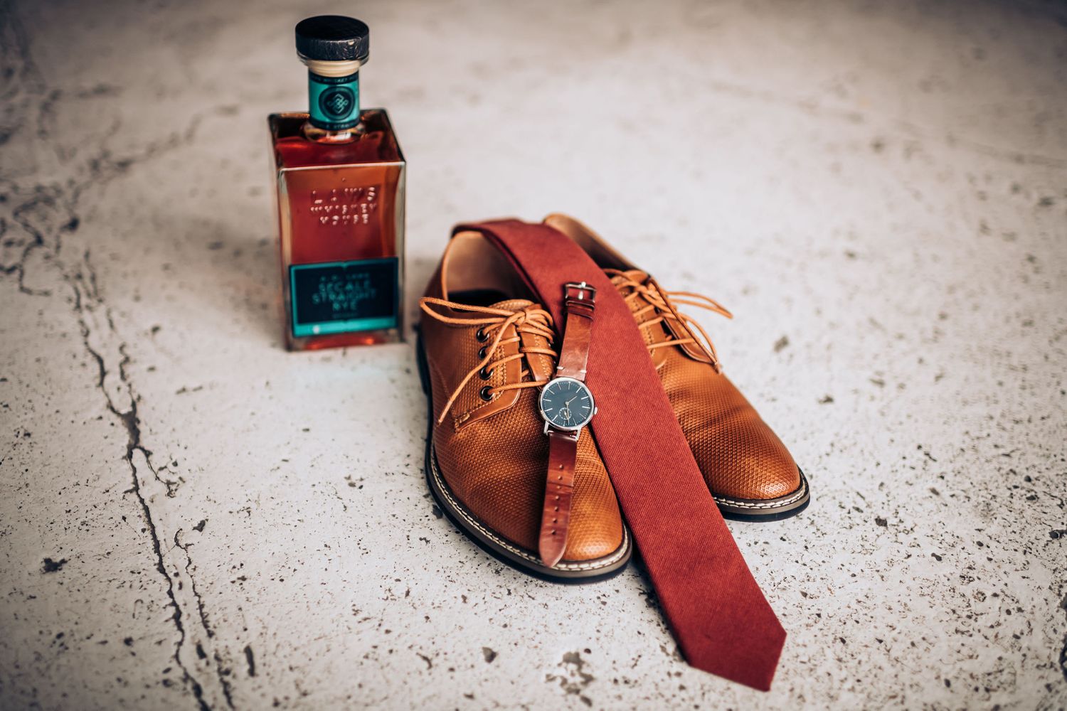 Groom's accessories including brown leather shoes, red tie and cologne bottle on concrete floor.