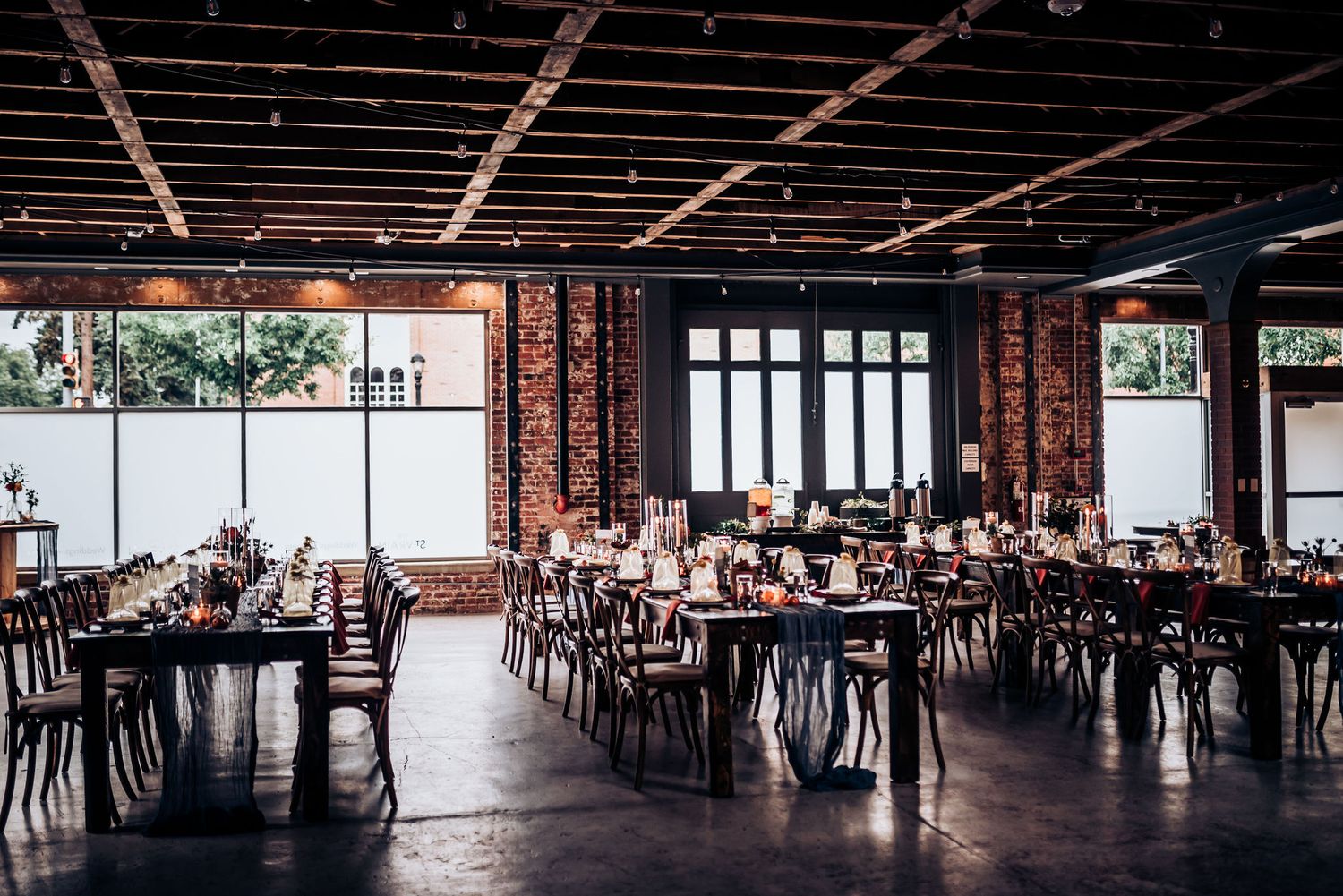 Spacious industrial wedding venue with large windows, exposed brick, and wooden cross-back chairs.