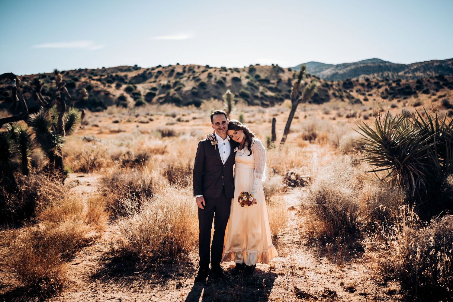 A wedding couple stands together in a desert landscape with rocky terrain and golden grass.