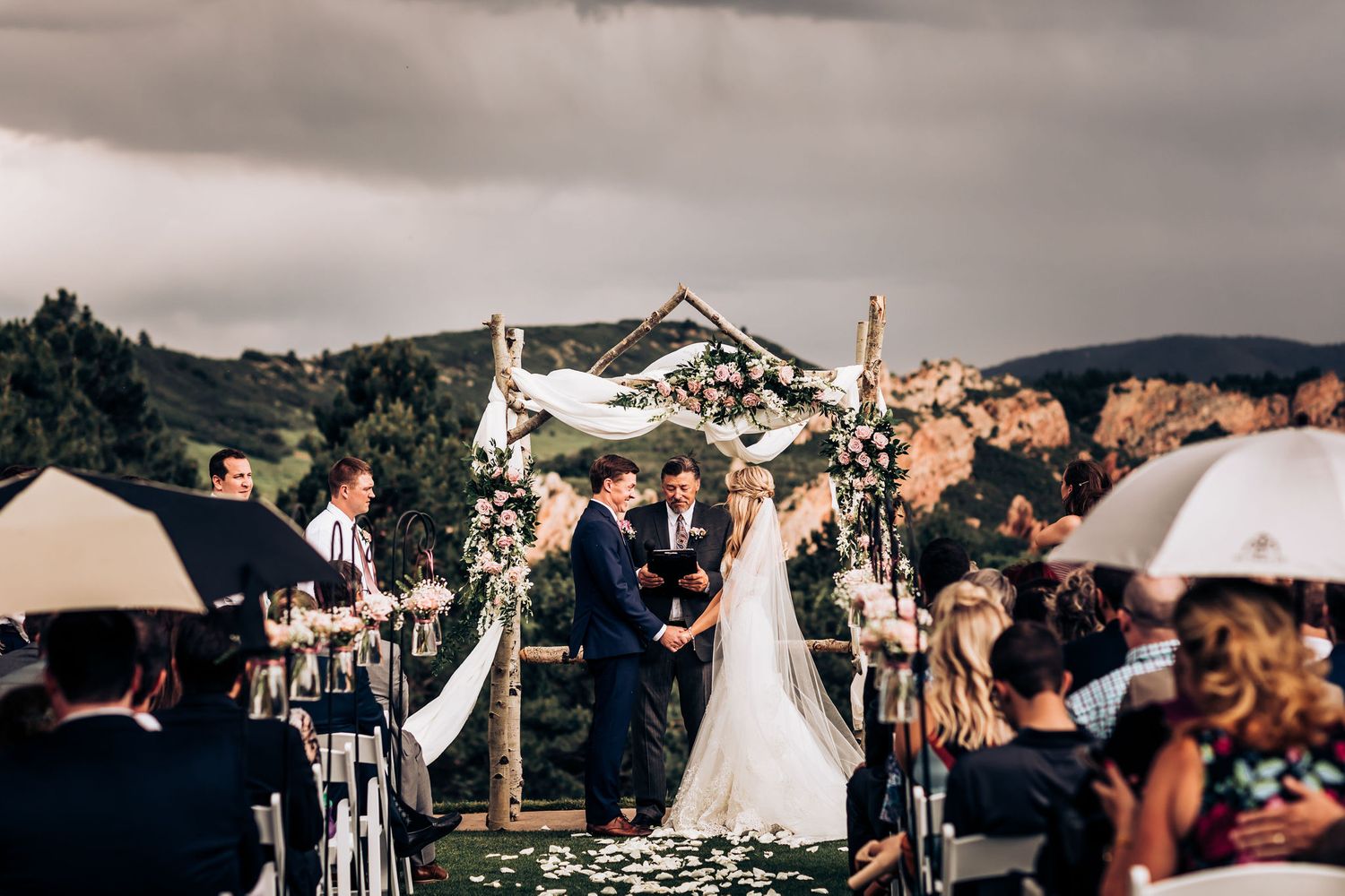 Outdoor wedding ceremony with white draped arch against cloudy mountain landscape.