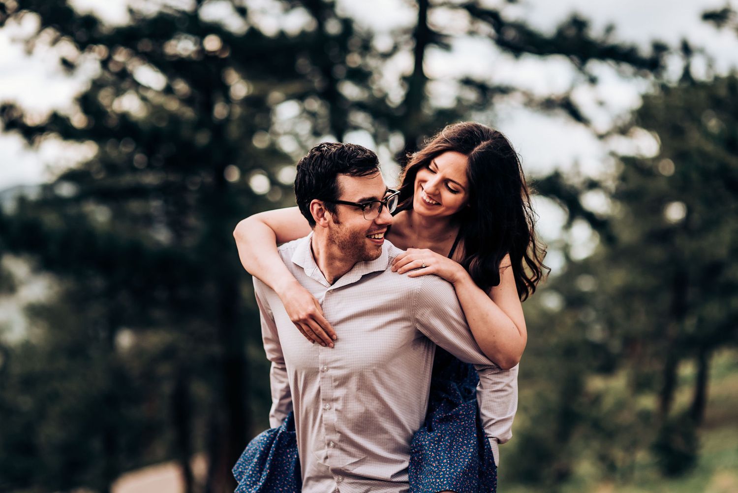 A playful couple shares a piggyback ride moment in a wooded outdoor setting.
