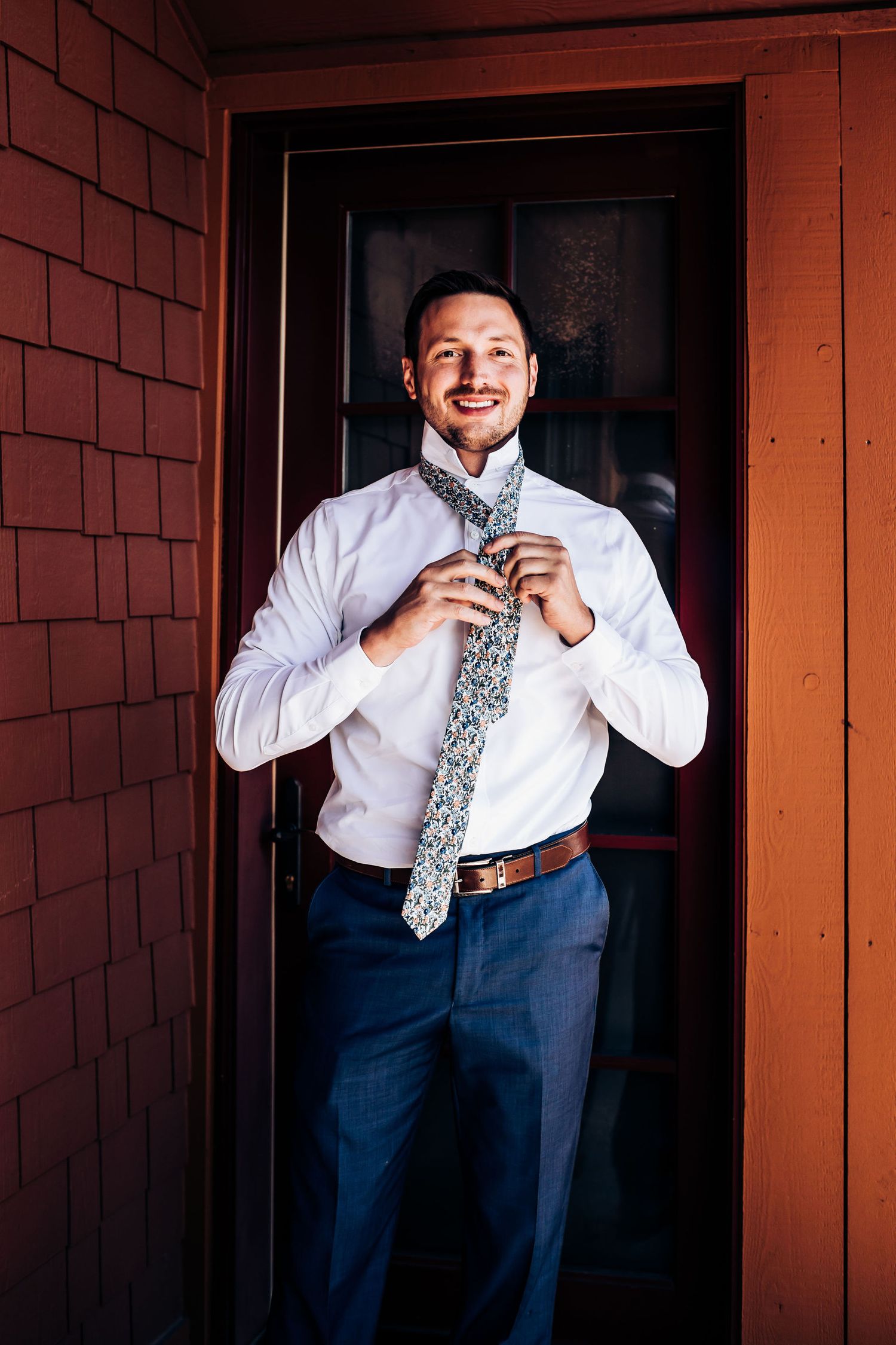A person adjusts a patterned gray tie while wearing a crisp white dress shirt and navy pants in front of an orange wall.