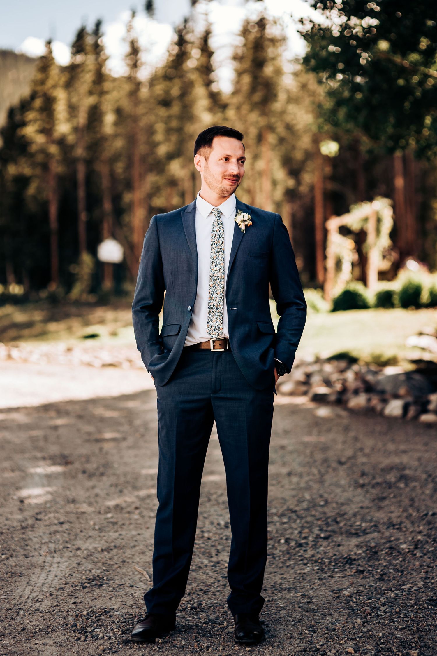 Groom in tailored navy suit poses confidently against forest background.