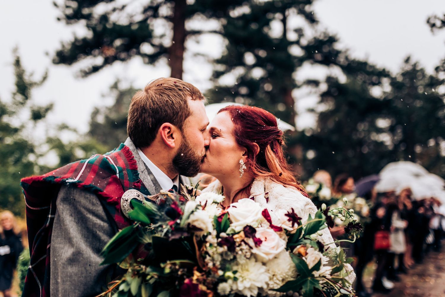 Romantic kiss between newlyweds surrounded by pine trees and wedding guests with umbrellas.