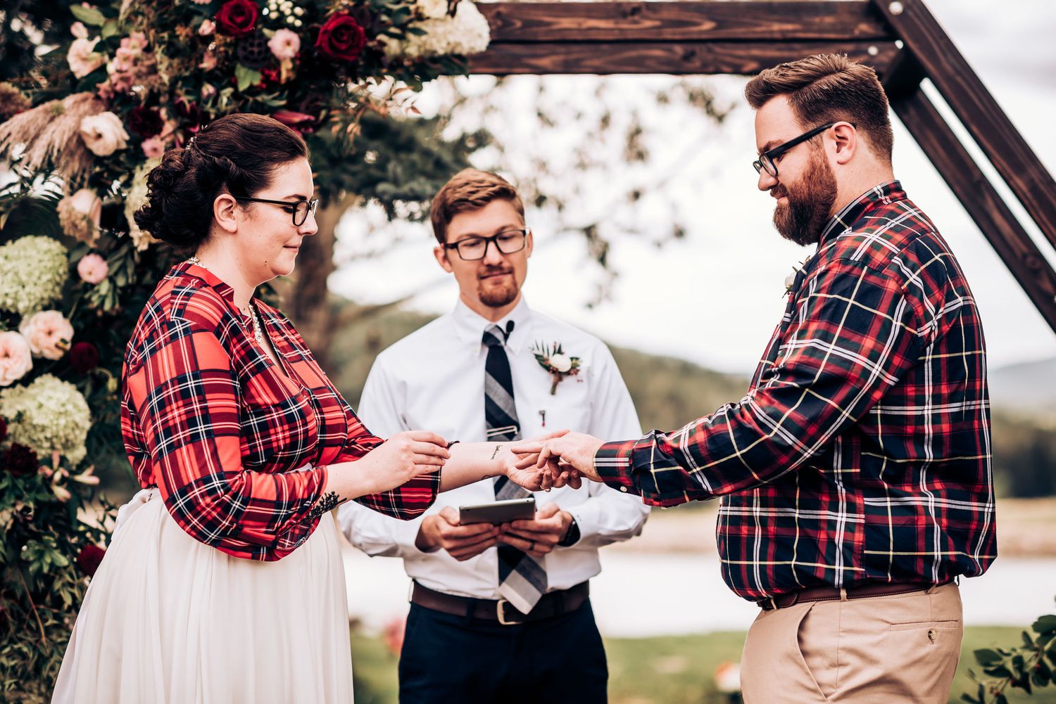 An outdoor wedding ceremony features a couple in matching plaid flannel exchanging vows with an officiant.