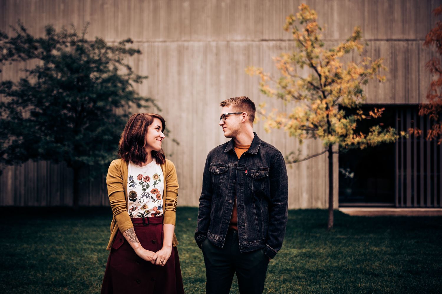 A couple poses outdoors with autumn trees and a wooden wall in the background.