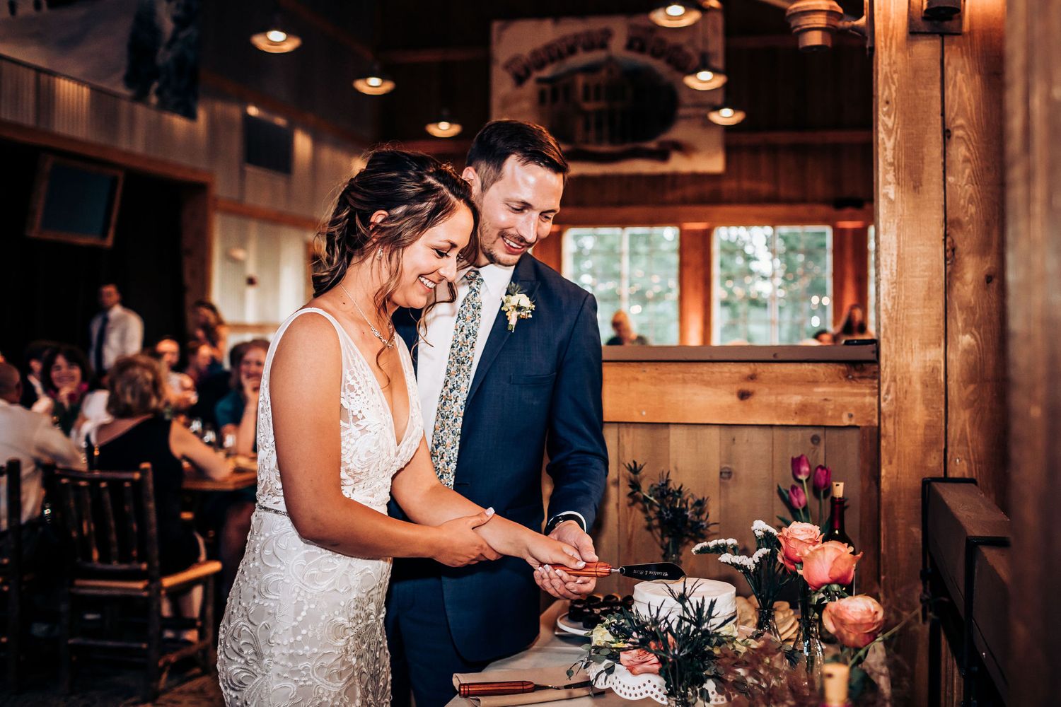 Couple cuts their wedding cake together in a rustic wooden reception venue.