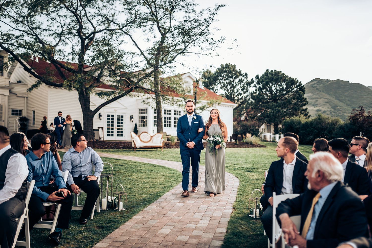 Wedding ceremony processional on stone pathway with seated guests and white colonial building backdrop.
