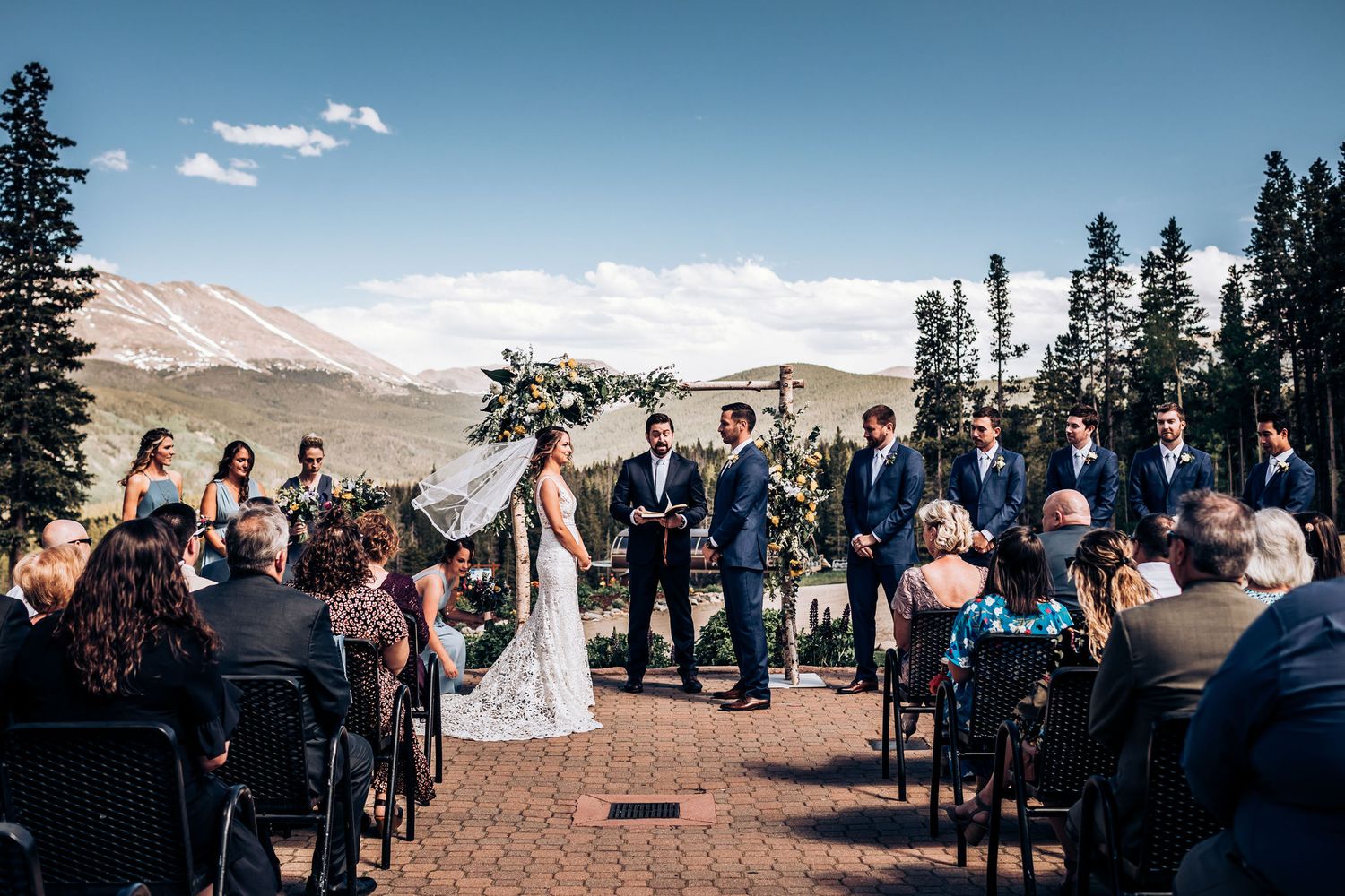 Outdoor mountain wedding ceremony with guests seated in rows facing a floral arch against a scenic backdrop.