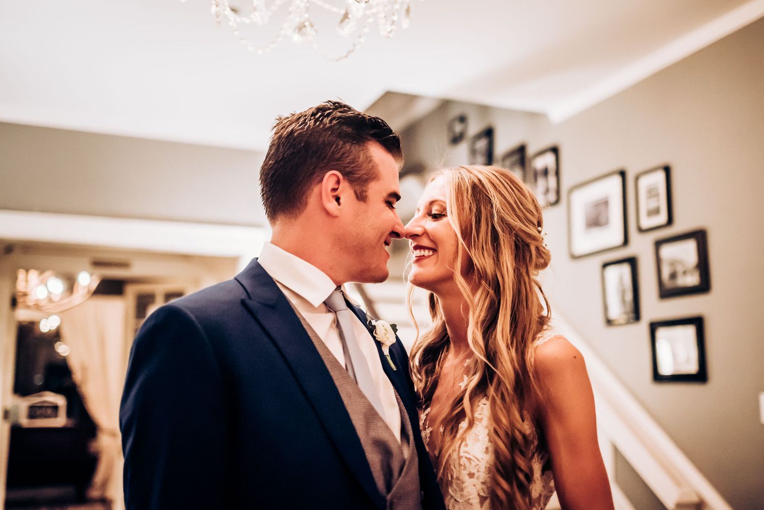 A romantic wedding moment captured indoors with elegant decor and framed photos in the background.