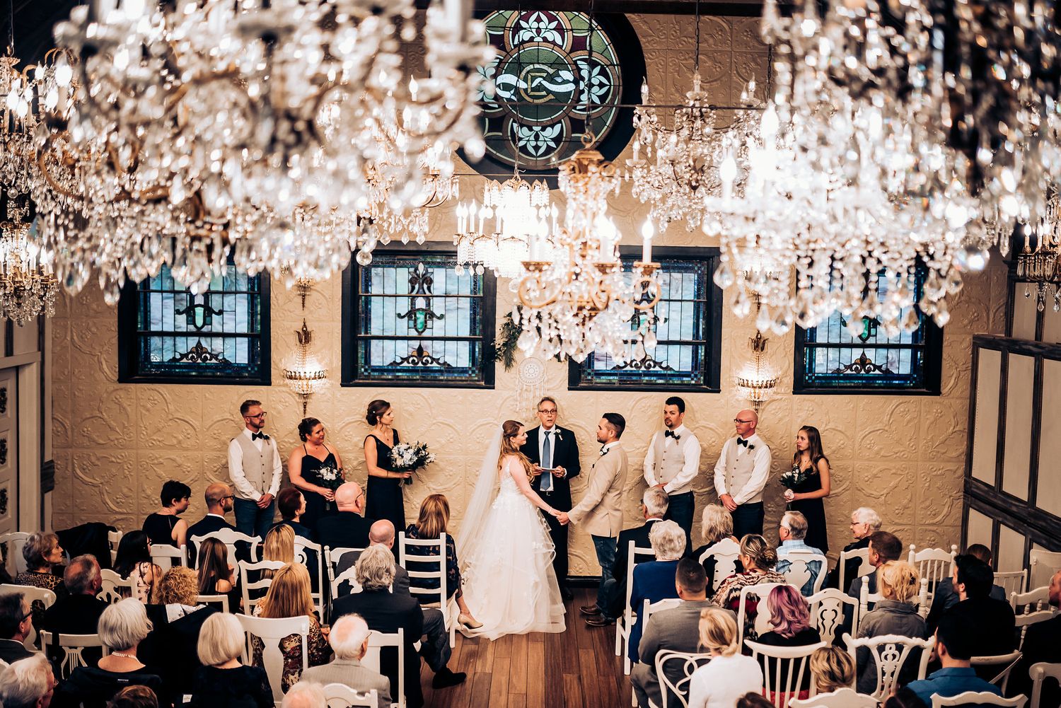Wedding ceremony viewed from above with crystal chandeliers and guests seated in white chairs.