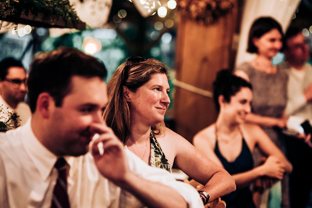Wedding guests smile and laugh while seated at reception tables with string lights decorating the venue.