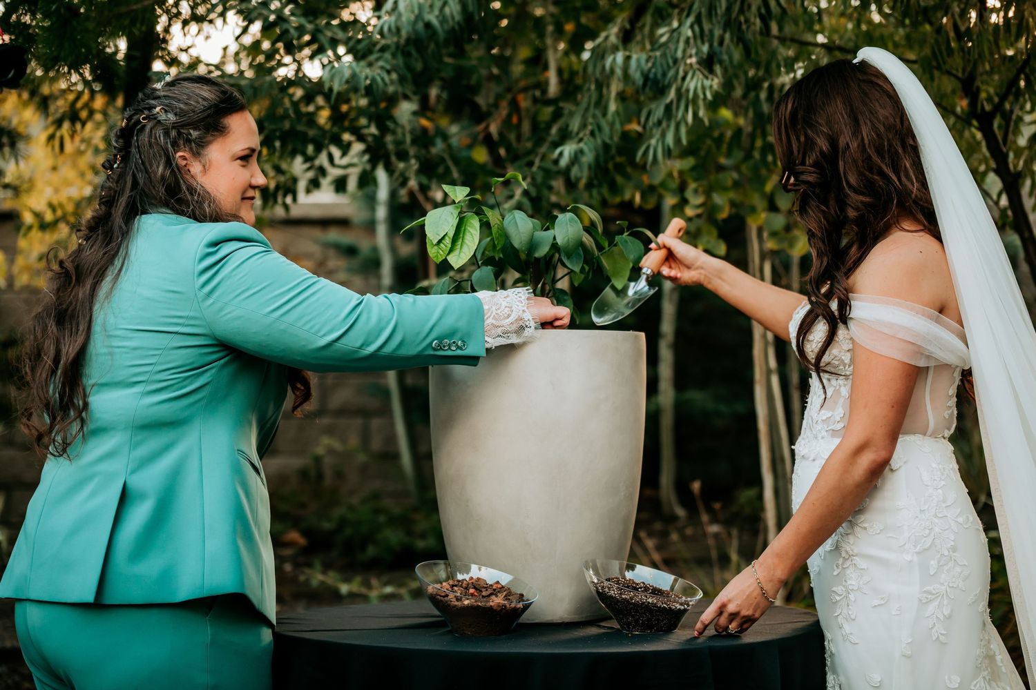 Smiling brides tend to a potted plant together during a symbolic tree planting wedding ceremony.
