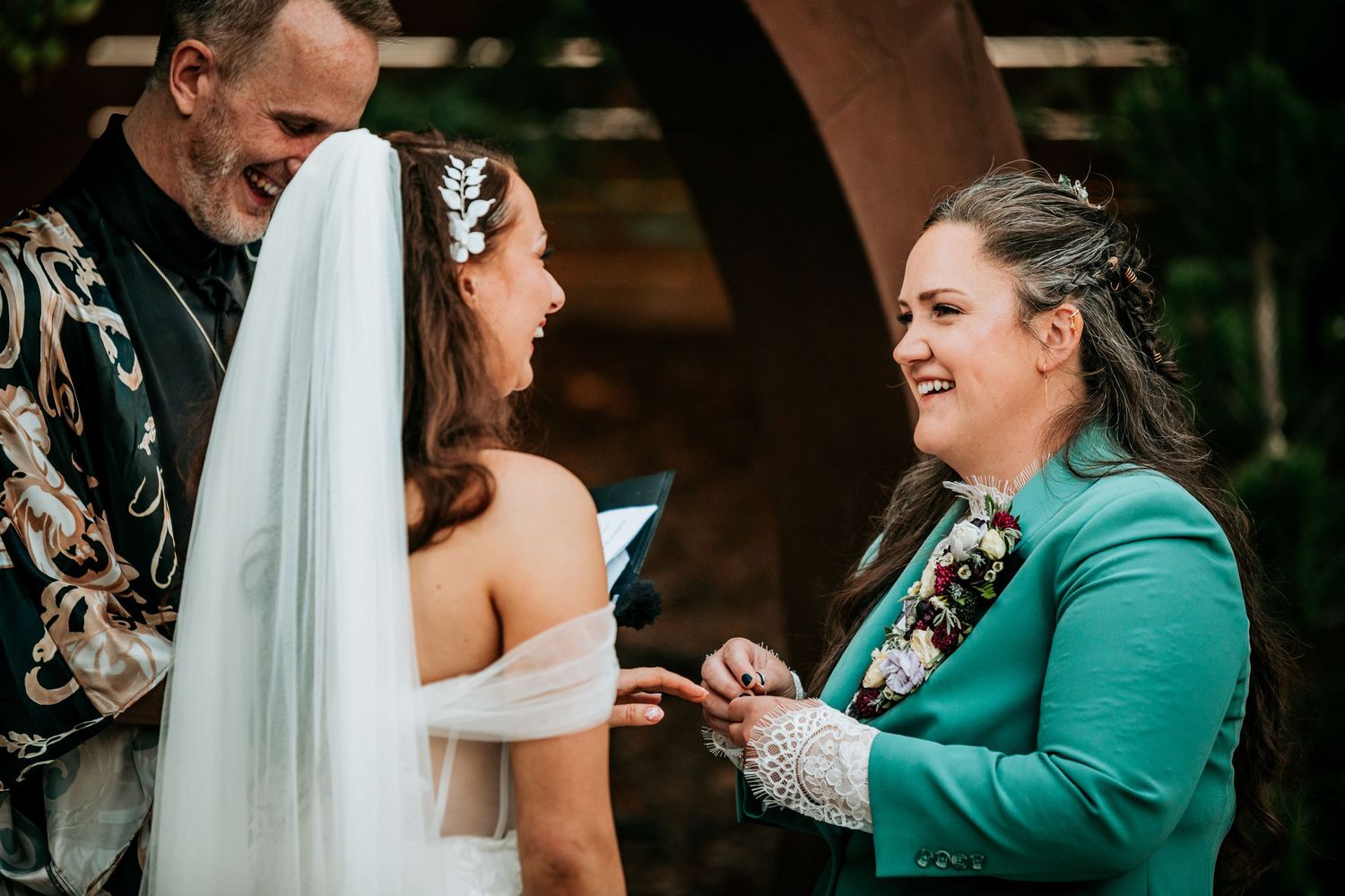 Two brides share a joyful moment during ring exchange ceremony with officiant in background.