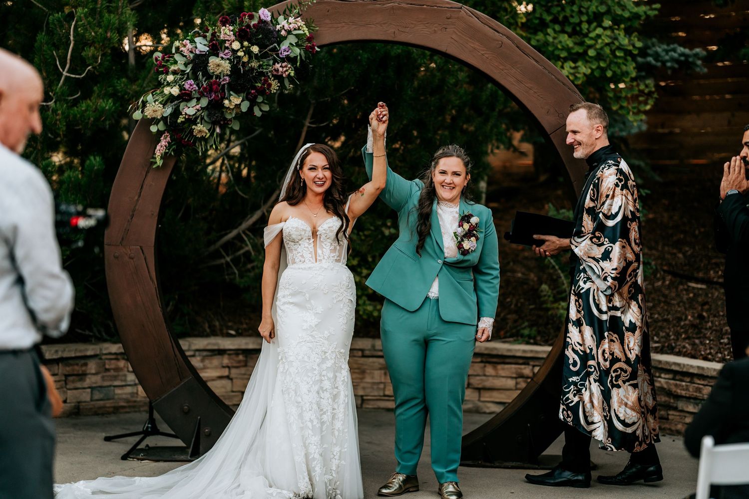 Two brides celebrate with raised hands as officiant cheers after outdoor wedding ceremony.