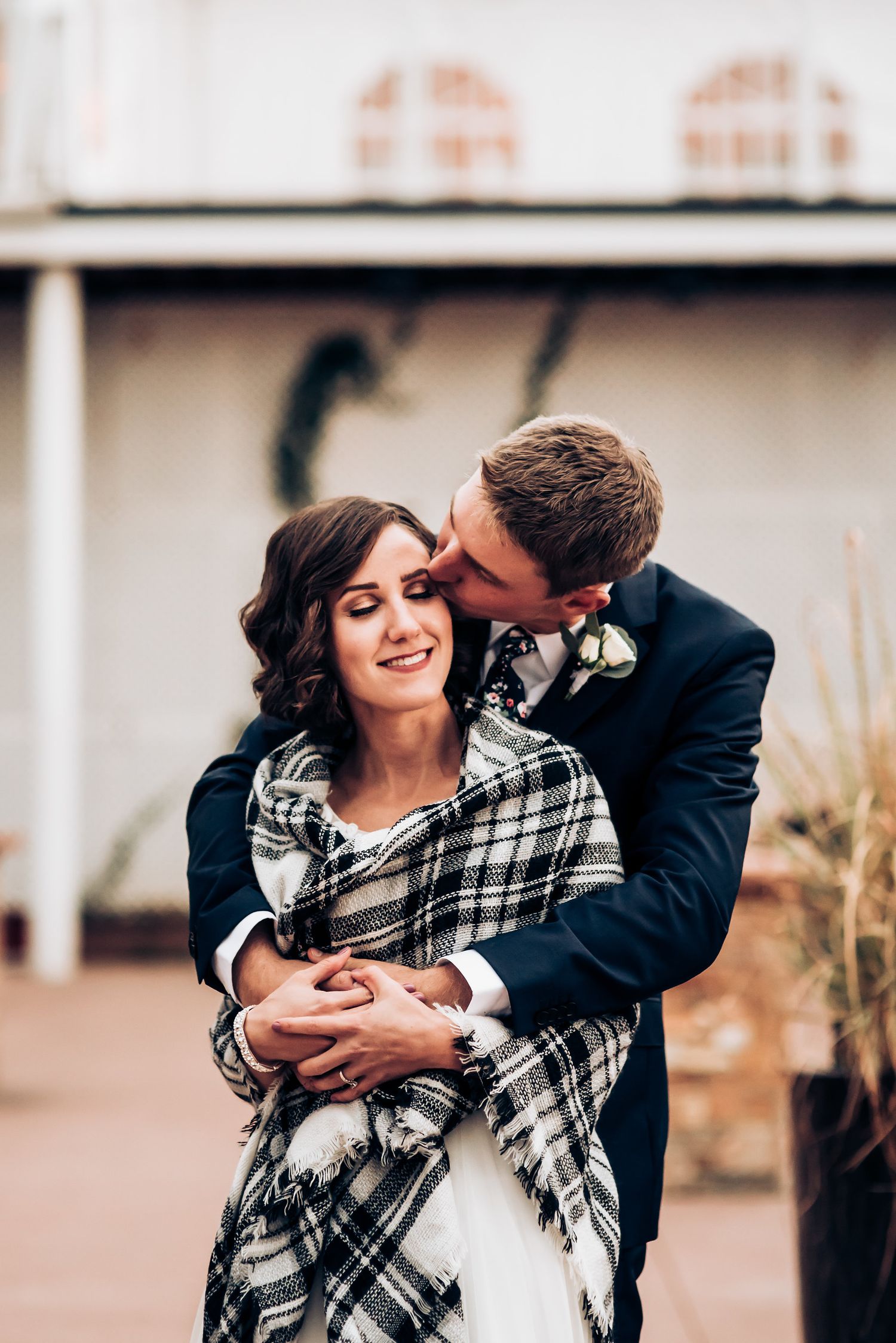 A couple shares an intimate moment while wrapped in a plaid blanket during their wedding photoshoot.