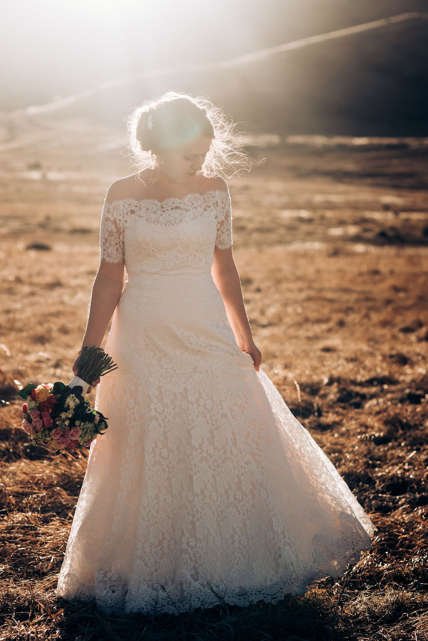 A bride in a lace wedding dress stands backlit against mountains with sunlight creating a glowing halo effect.
