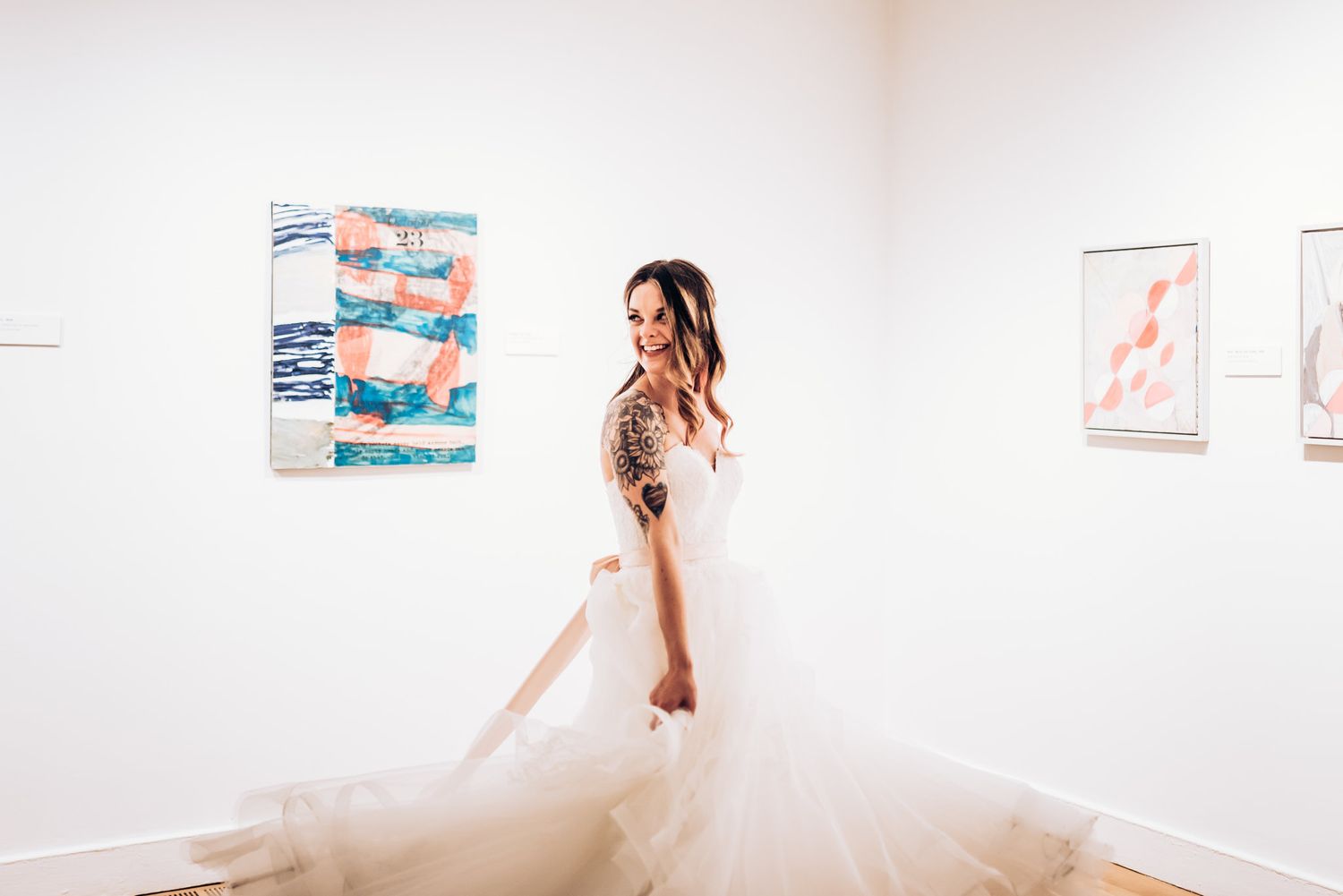 A bride in a flowing white dress poses in an art gallery with abstract paintings.