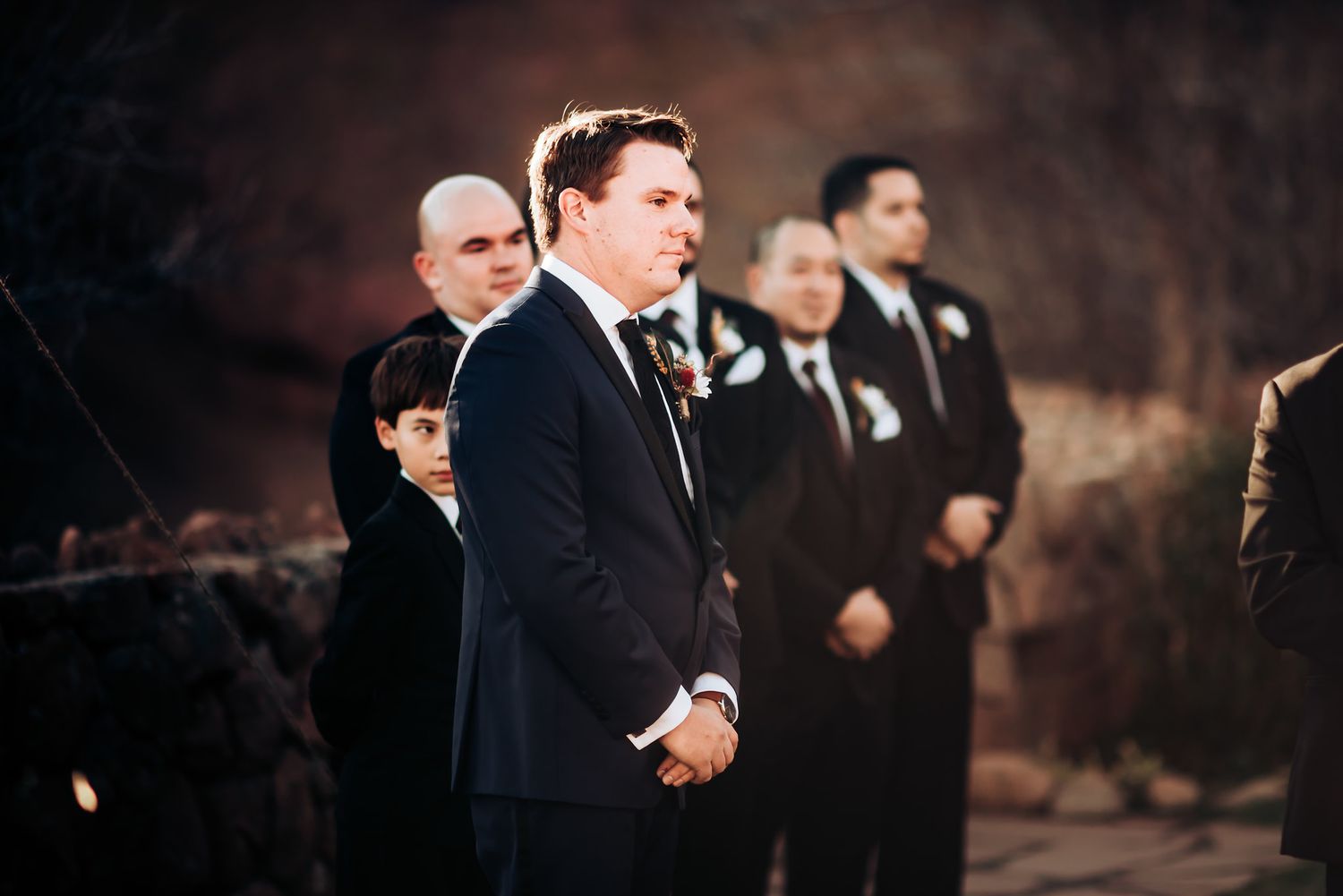 Wedding party members in formal attire stand in line during outdoor ceremony.