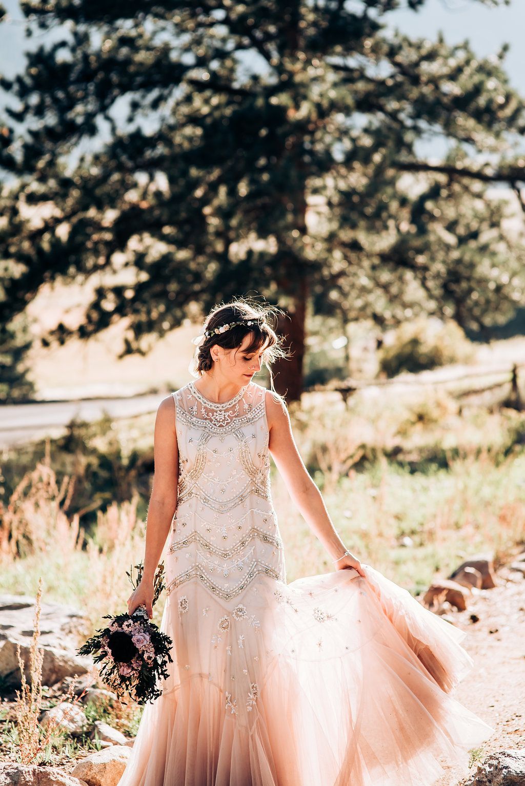 Blush beaded wedding gown flowing in mountain breeze with pine trees.