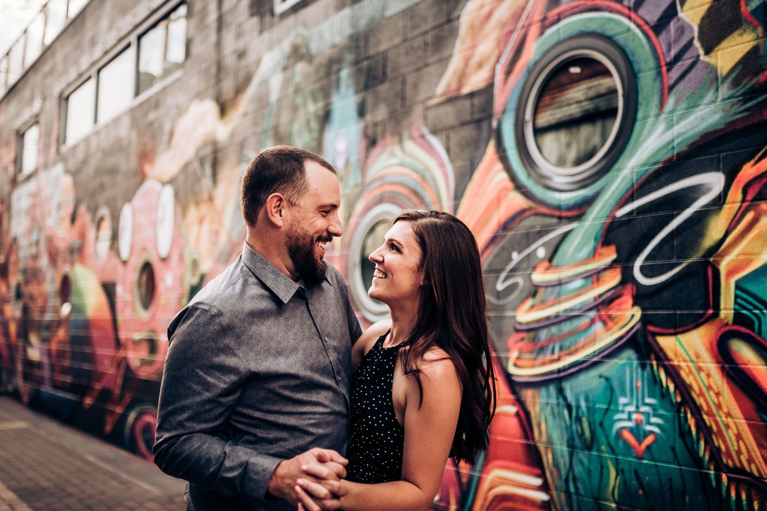 A couple laughs together in front of an urban mural with teal circular patterns.