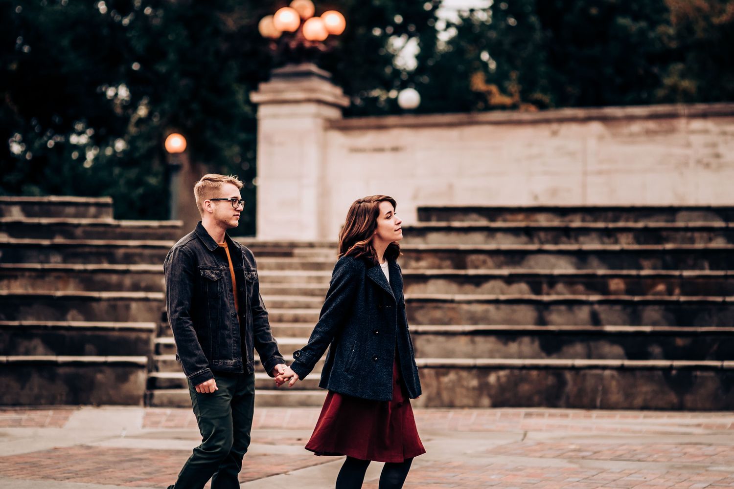 A romantic couple walks hand in hand near historic stone steps with glowing lamps in background.