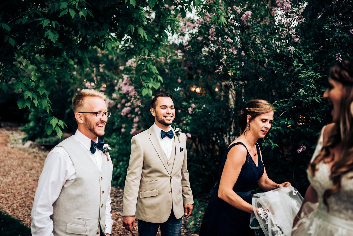 Wedding party members gather outdoors among lush green foliage wearing formal attire.