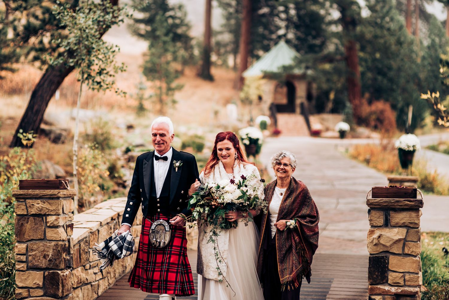 Three people walk down a stone path at a wedding, with the center person wearing a white bridal gown.
