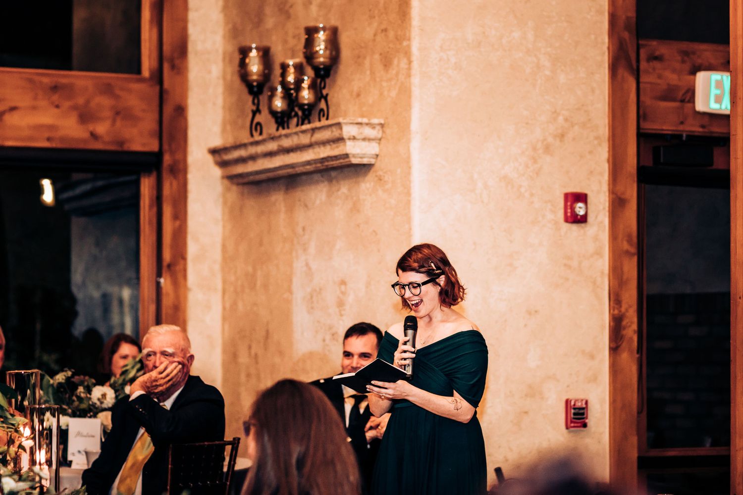 Guest stands to make a speech during an intimate wedding reception in a warm-lit venue.
