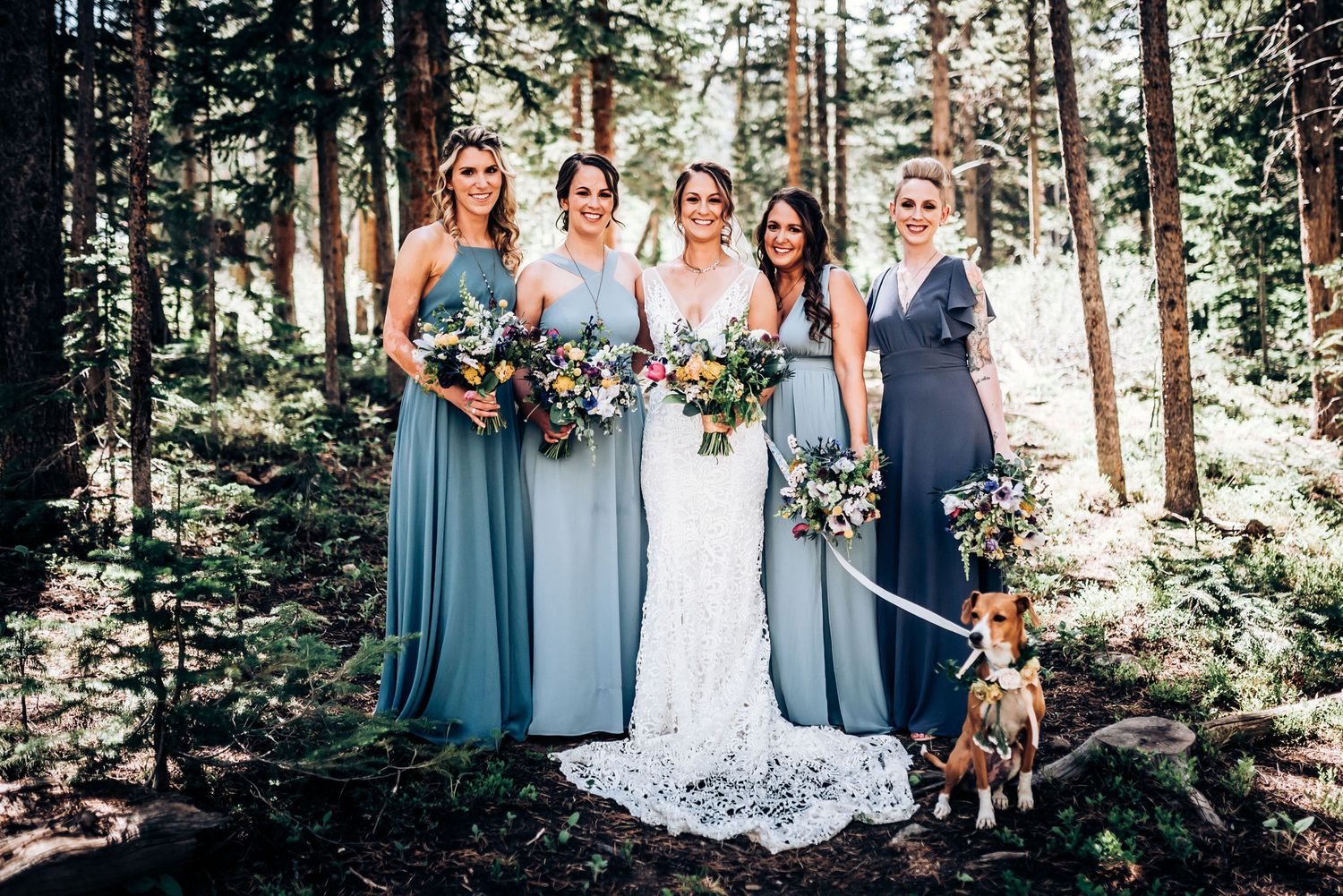 Bridal party poses in forest setting with dog while holding colorful bouquets.