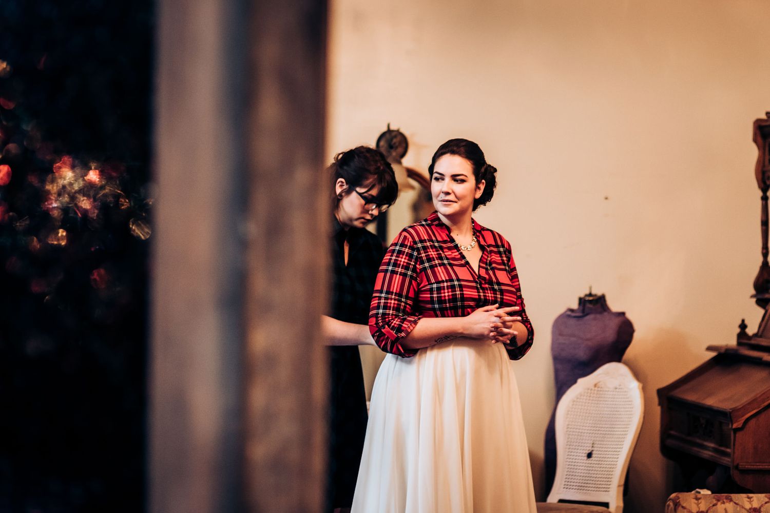 A person in a red plaid shirt and white skirt stands in a dimly lit room near a dress form.