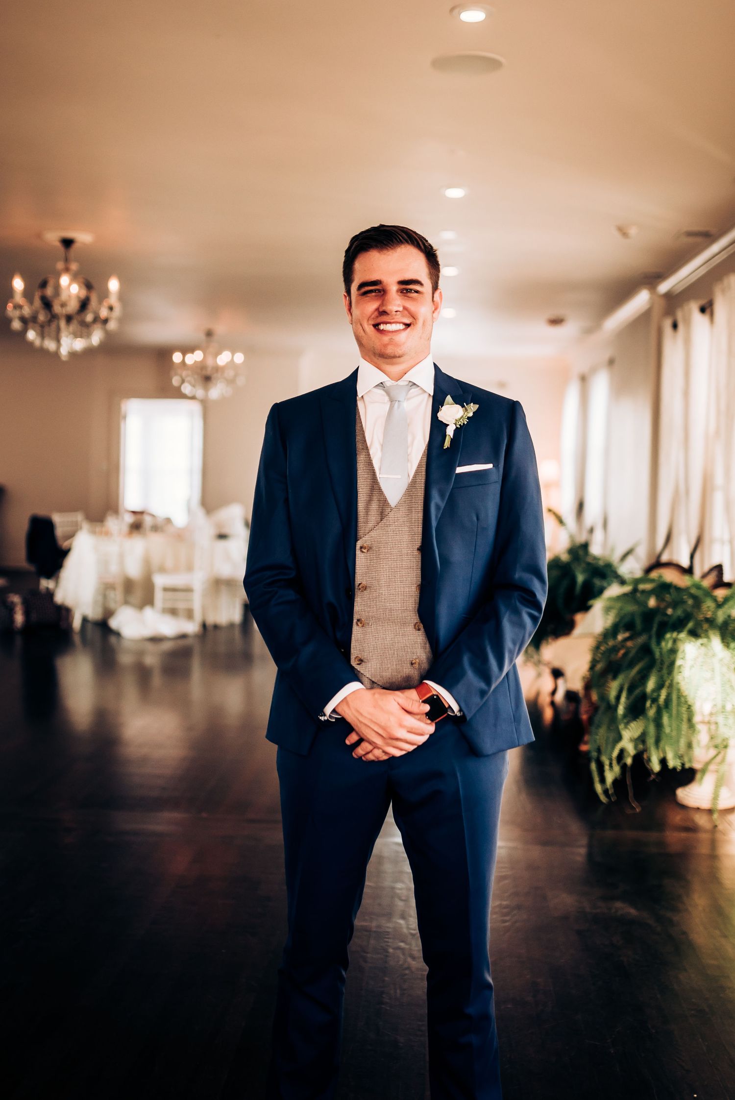 A groom in a navy suit with tan vest stands in an elegant venue with chandeliers and white curtains.