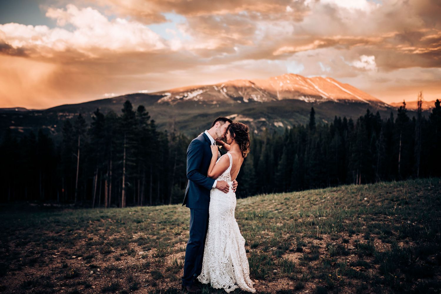 Wedding couple shares romantic moment on mountainside with dramatic orange sky.