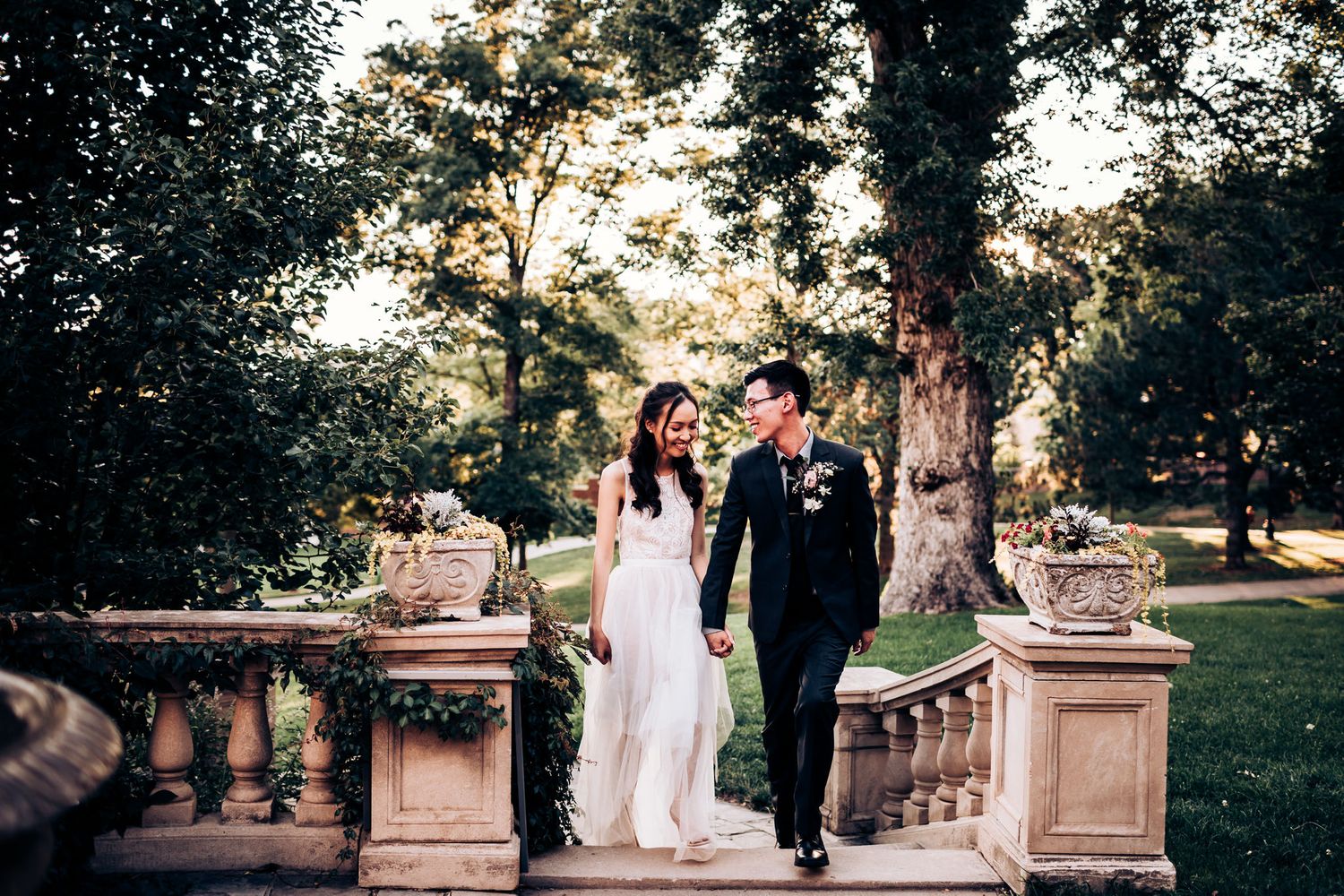 A couple walks down ornate stone steps in a romantic garden setting at golden hour.