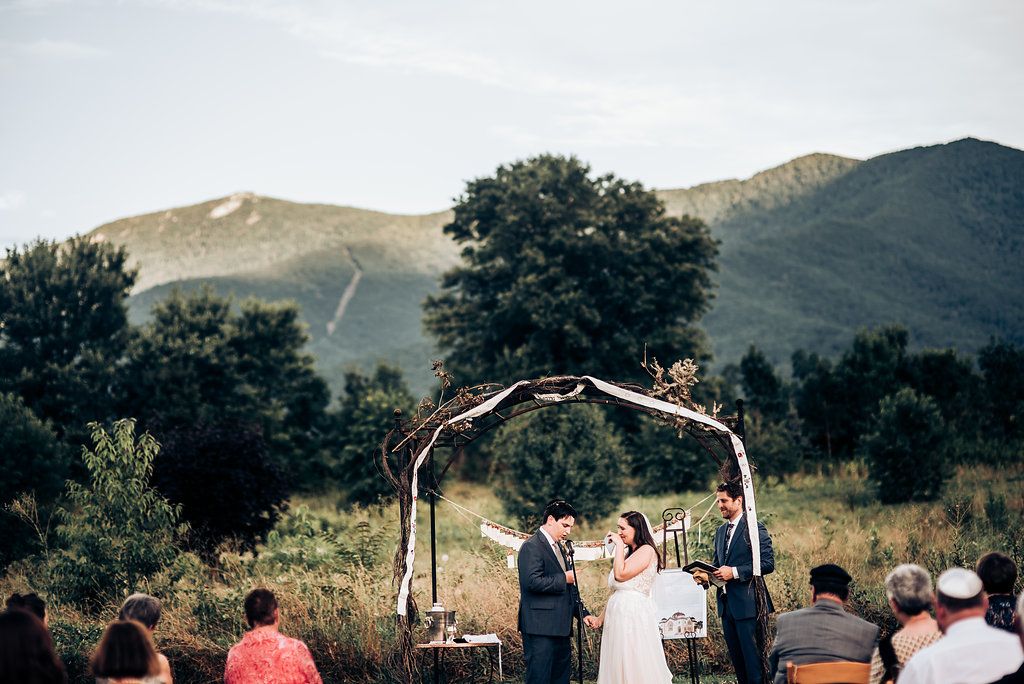 Wedding ceremony taking place under decorated arch with mountain landscape in background.