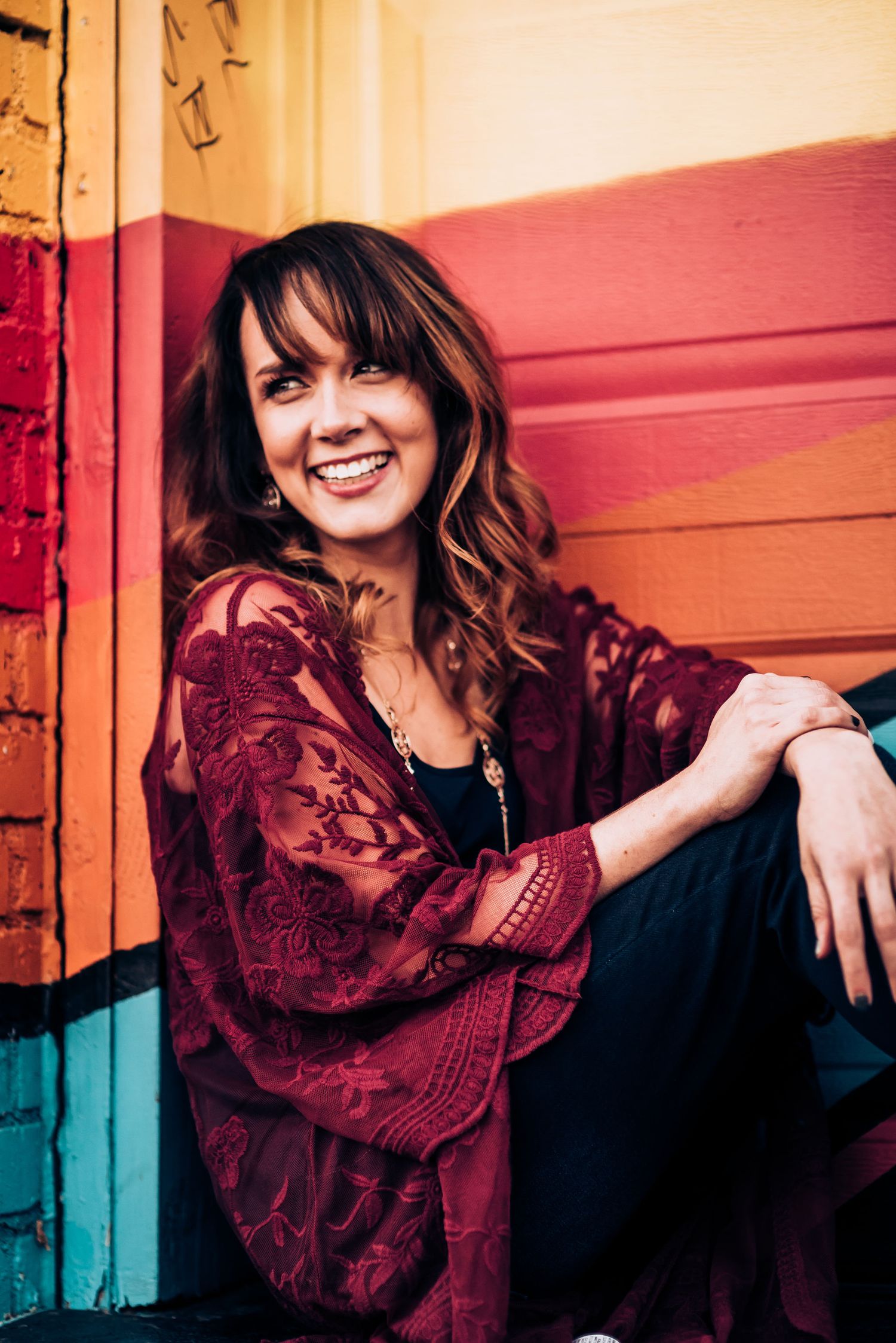 Woman in burgundy lace top smiles brightly against a colorful striped wall.