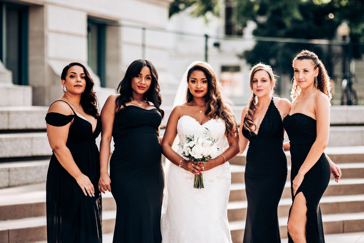 A bridal party in elegant black dresses poses on stone steps outdoors.