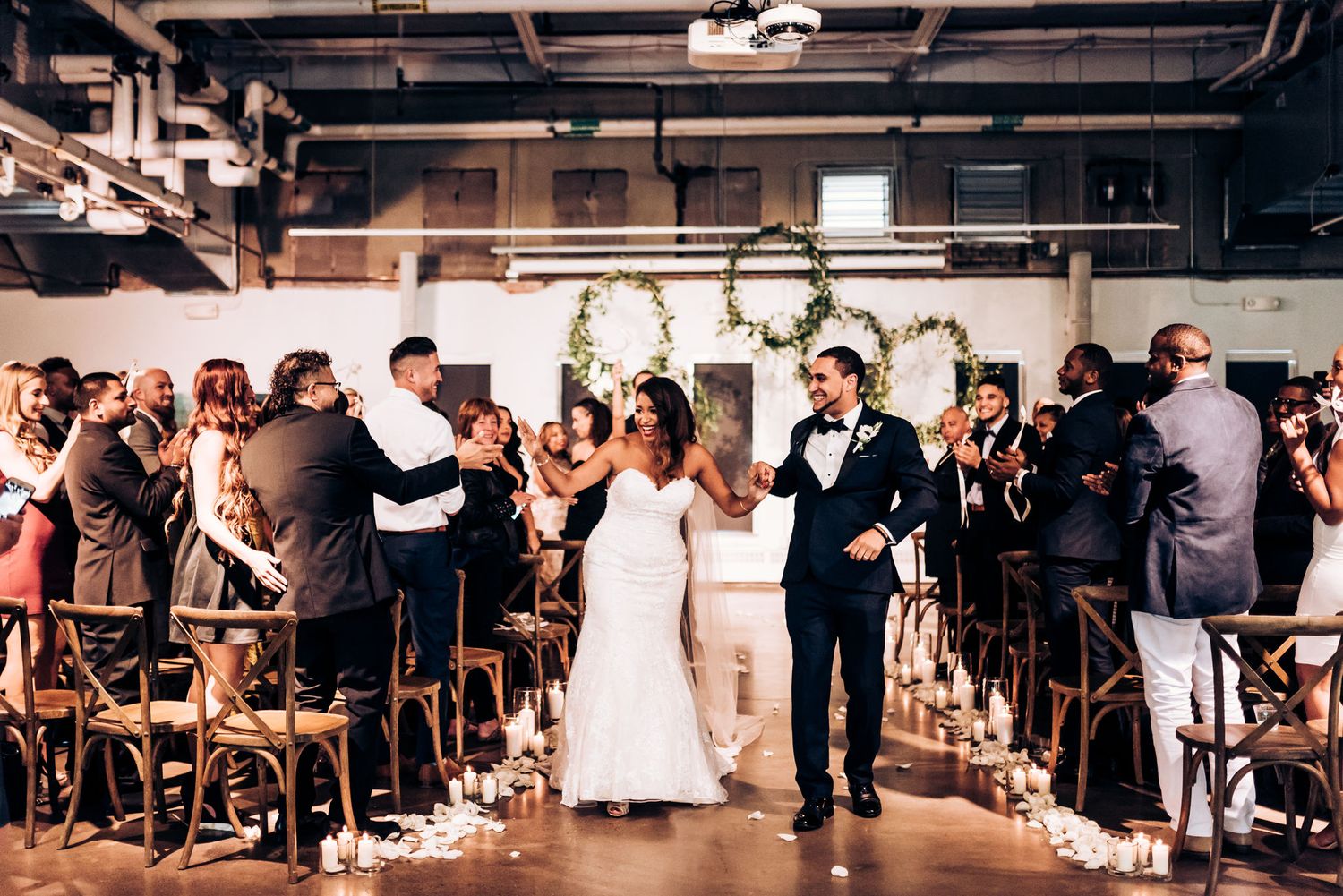 Wedding party exits down aisle decorated with candles in modern industrial reception space.
