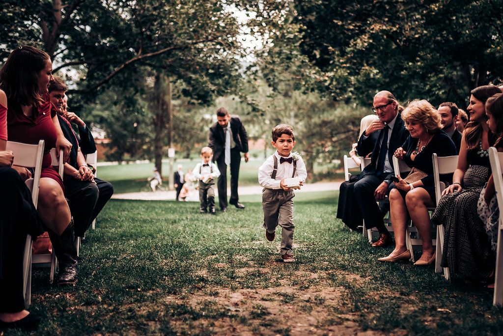 Young ring bearer in suspenders walks down outdoor wedding aisle between rows of seated guests.