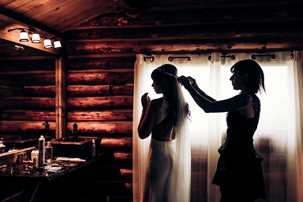 Silhouettes prepare for a wedding in rustic log cabin with warm lighting and window backdrop.