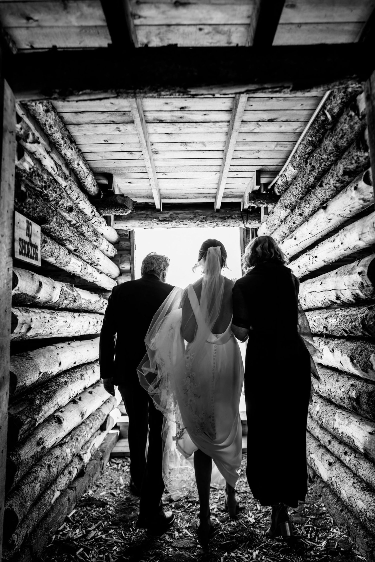 Black and white silhouettes of three people walking through rustic log cabin corridor toward bright light.