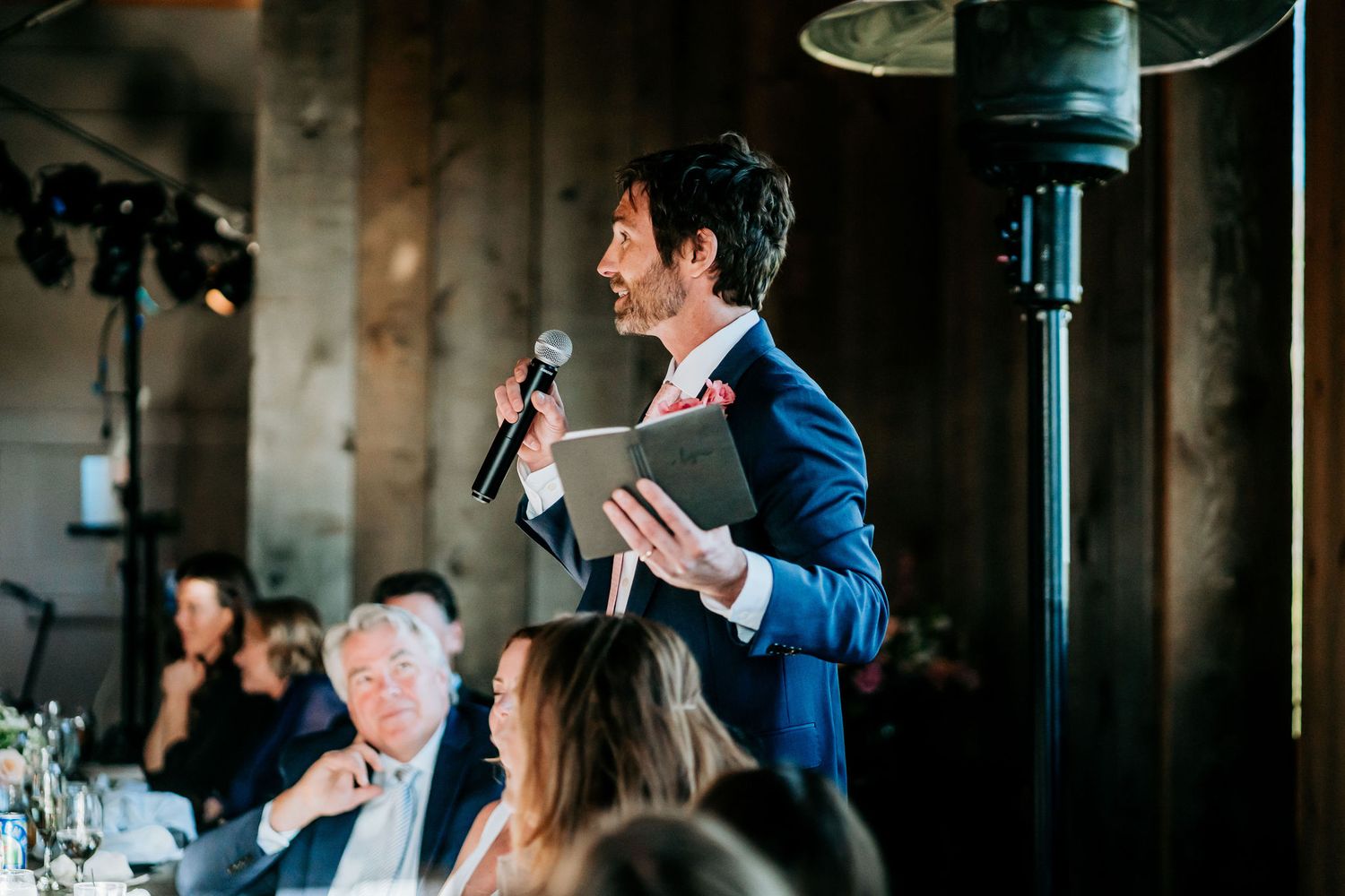 A speaker in a blue suit addresses wedding guests during dinner reception.